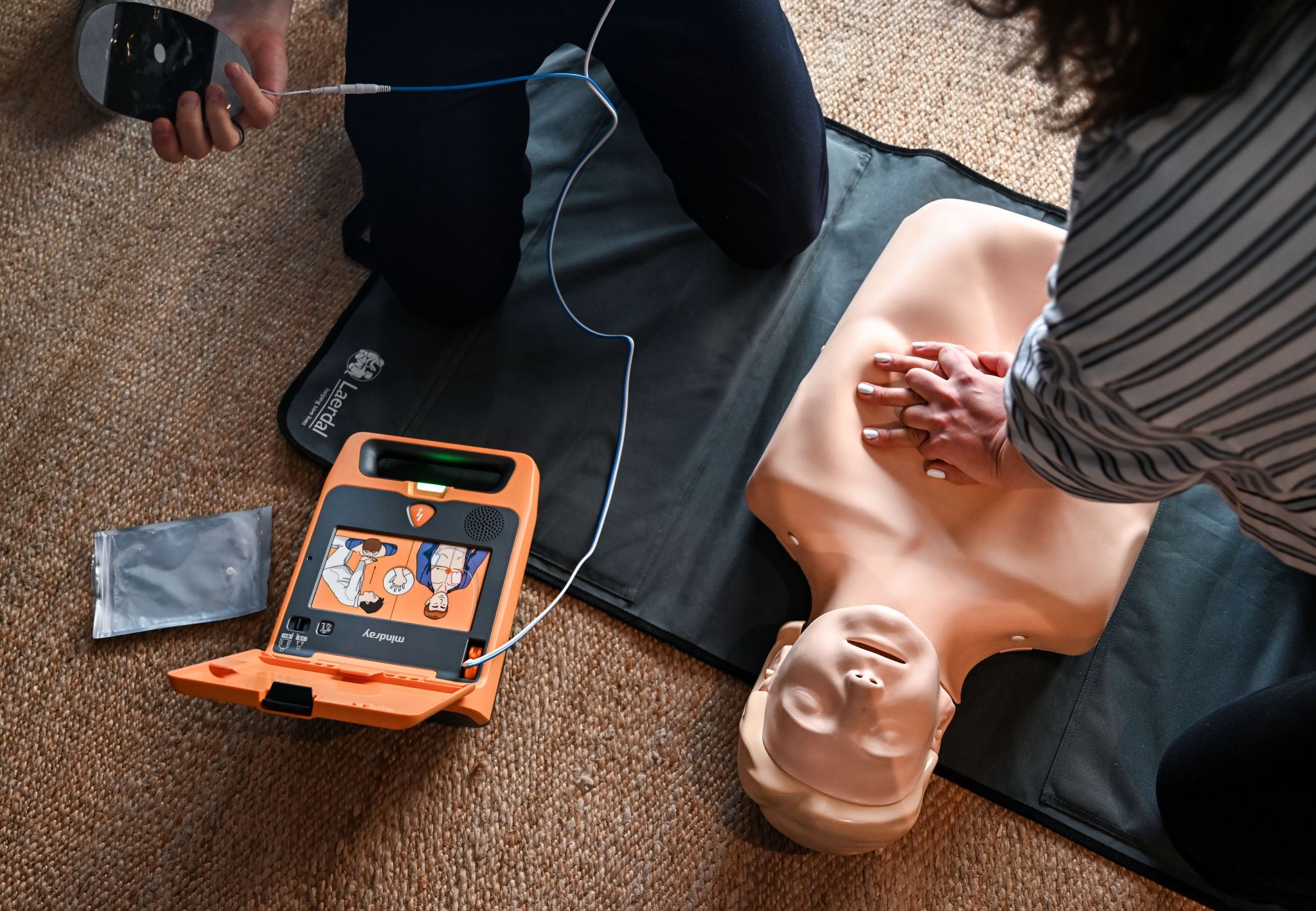 Reanimation mit dem Mindray BeneHeart AED-Defibrillator: Helfer führt eine Herzdruckmassage durch, während das geöffnete orange Defibrillator-Gerät mit Elektroden bereitsteht.